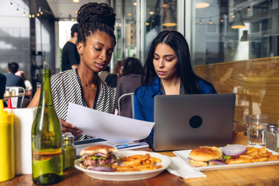 Two women review paperwork at an outdoor cafe table, with burgers and fries in front of them, an open laptop, and condiments and a green bottle nearby.