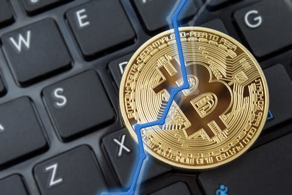 A rising blue line overlaid on a physical gold bitcoin, with a black keyboard in the background.