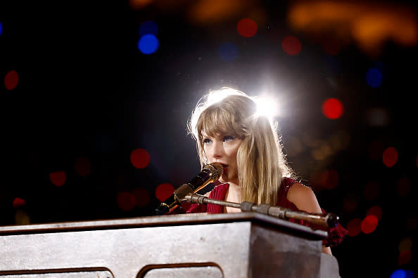 Taylor Swift performing at a microphone with a sparkling backdrop. She wears a sleeveless top