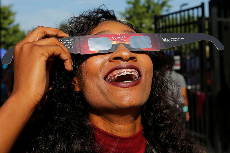 A cheerleader smiles as she looks up at the sky with eclipse glasses in her hand.