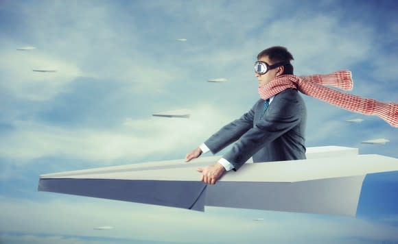 A man in a gray suit with a red scarf and goggles flies in a paper airplane.