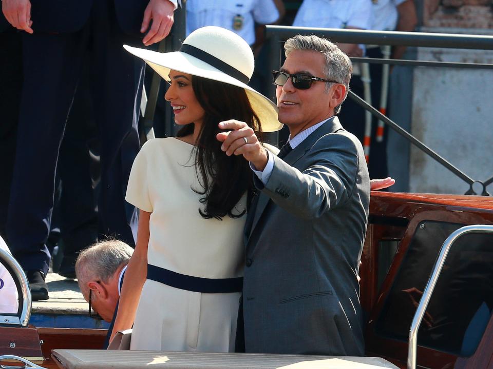 Amal Clooney in a white suit with a large, white floppy hat and George Clooney in a gray suit