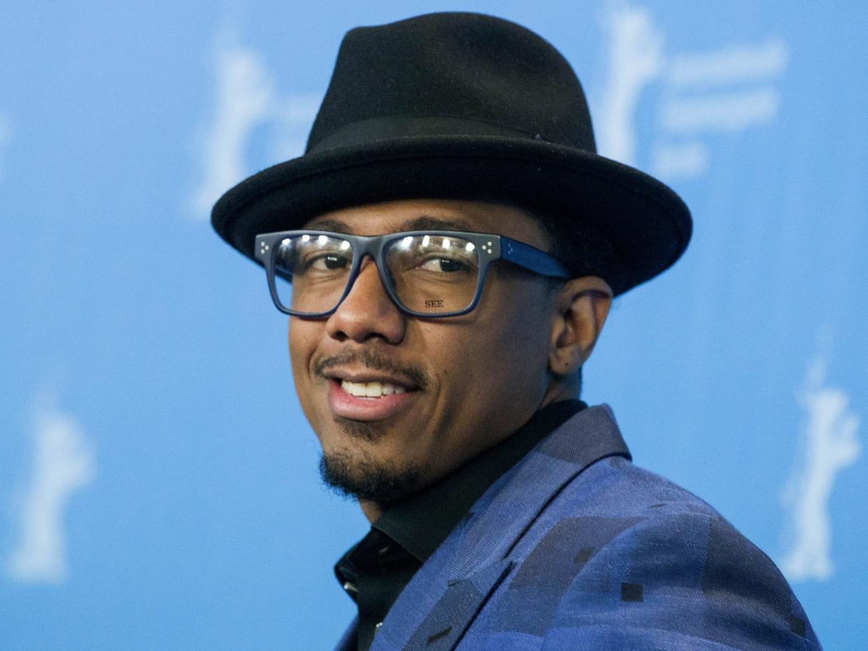 Nick Cannon in a black hat, blue suit and glasses in front of a light blue backdrop