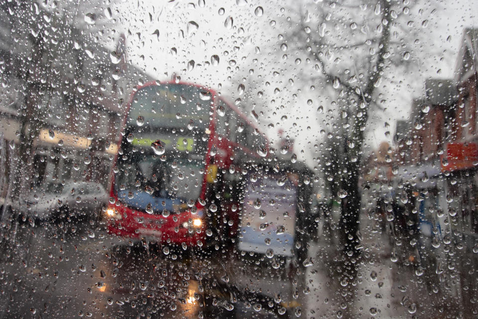 [UNVERIFIED CONTENT] Raindrops on a transparent surface, with an out-of-focus background. Awful british weather in this outdoor scene with torrential rain. A red London bus with its headlights on has stopped to collect passengers at a bus stop. A damp and grey british high street scene. London, UK