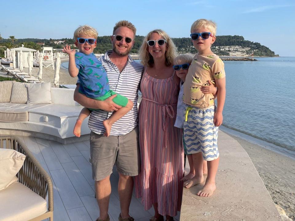 Photo of her Kat Storr, her husband, and three sons standing on an outdoor patio by the beach. Kat has her arms around her husband and one of her sons. She wears a red, blue, and white striped strappy dress and white sunglasses. Her husband has blonde hair spiked up with gel and wears sunglasses, a blue and white striped buttondown shirt, and grey shorts. He holds one son on his right hip. That son wears a blue t-shirt, sunglasses, and green shorts, and is waving at the camera. Two of the sons are standing on a low brick wall to Kat's right. The one closest to her wears a white t-shirt and sunglasses. His brother stands to this right and wears a tan shirt with blue and white striped shorts and sunglasses.