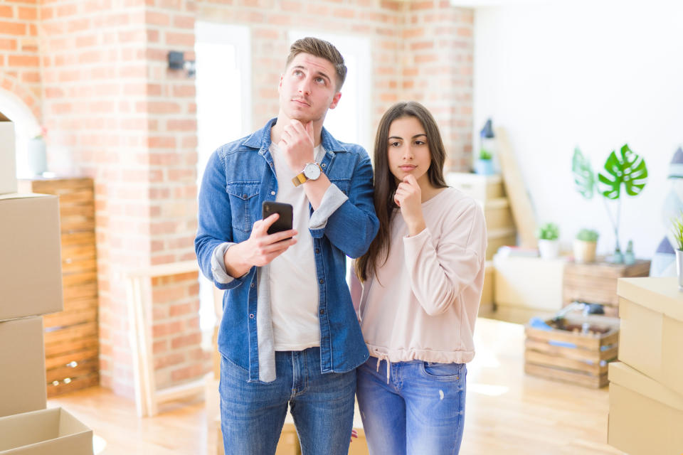 A young couple standing in their new house holding a smartphone and looking like they are thinking about something.