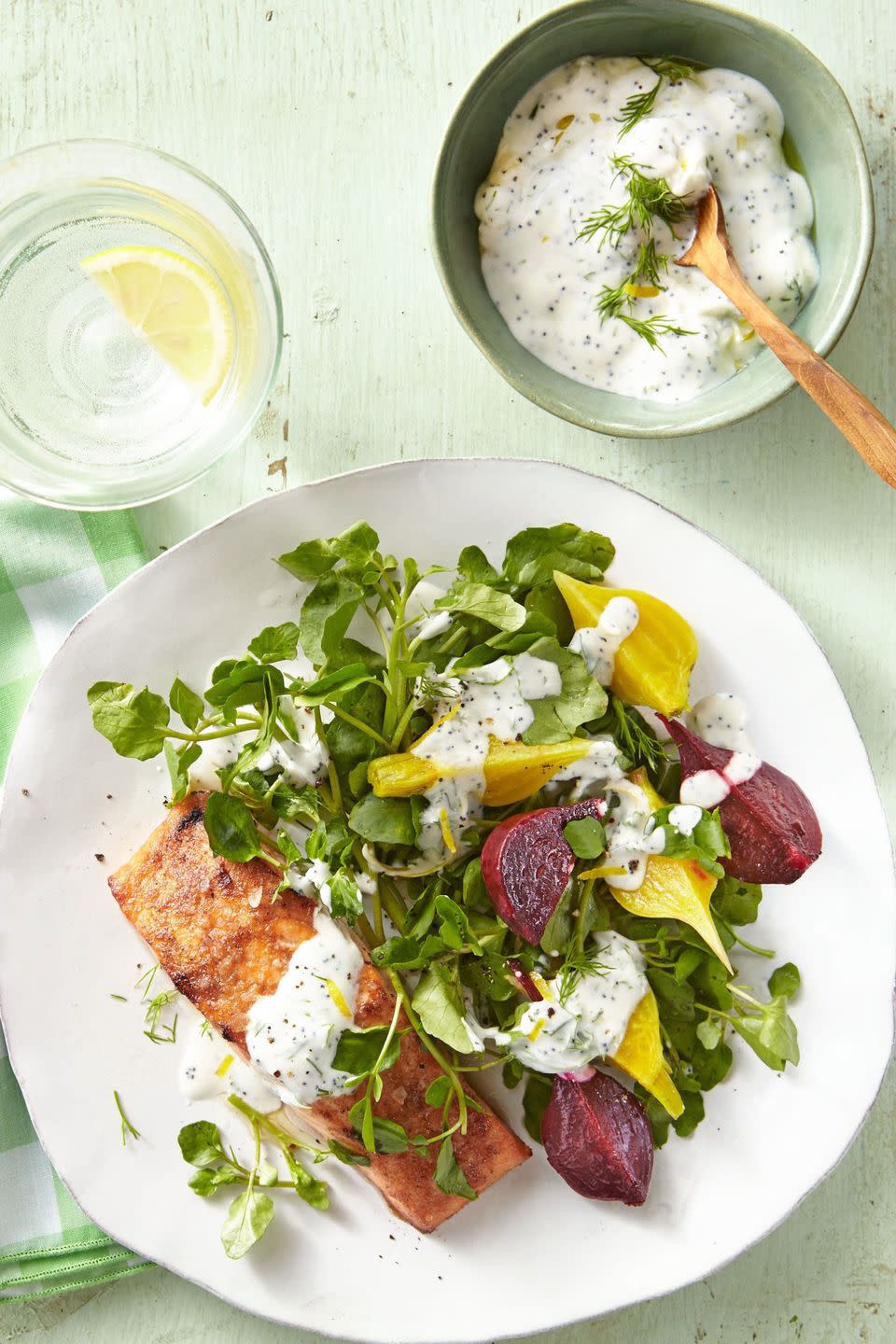 salmon and beets with yogurt sauce over watercress on a white plate with a glass of water with a lemon wedge and a small bowl of extra dressing