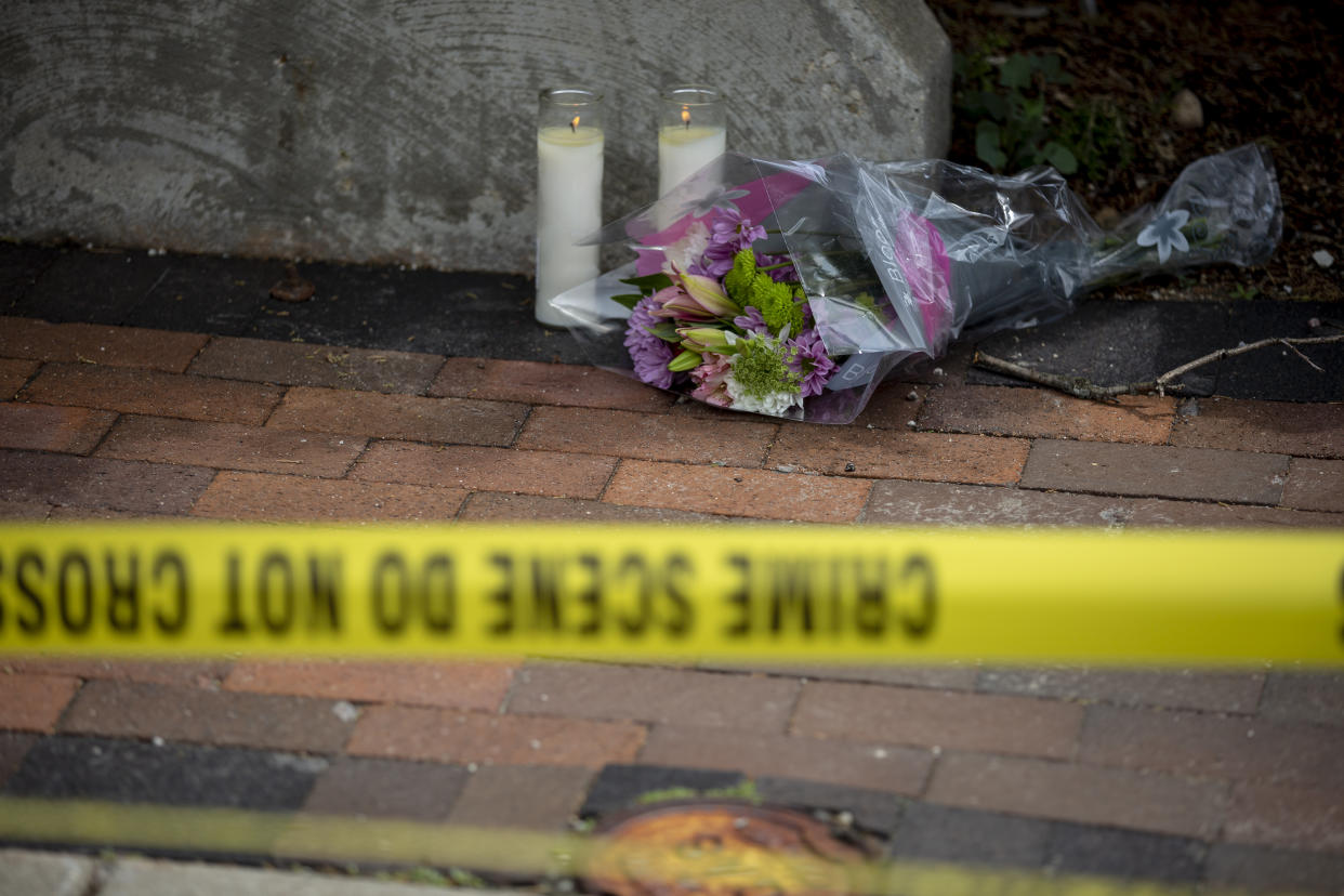 Two candles are lit on the sidewalk next to some flowers behind a yellow crime tape.