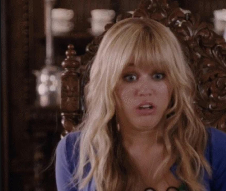 A surprised Hannah Montana, played by Miley Cyrus, sits on an ornate wooden chair with an expression of shock and concern