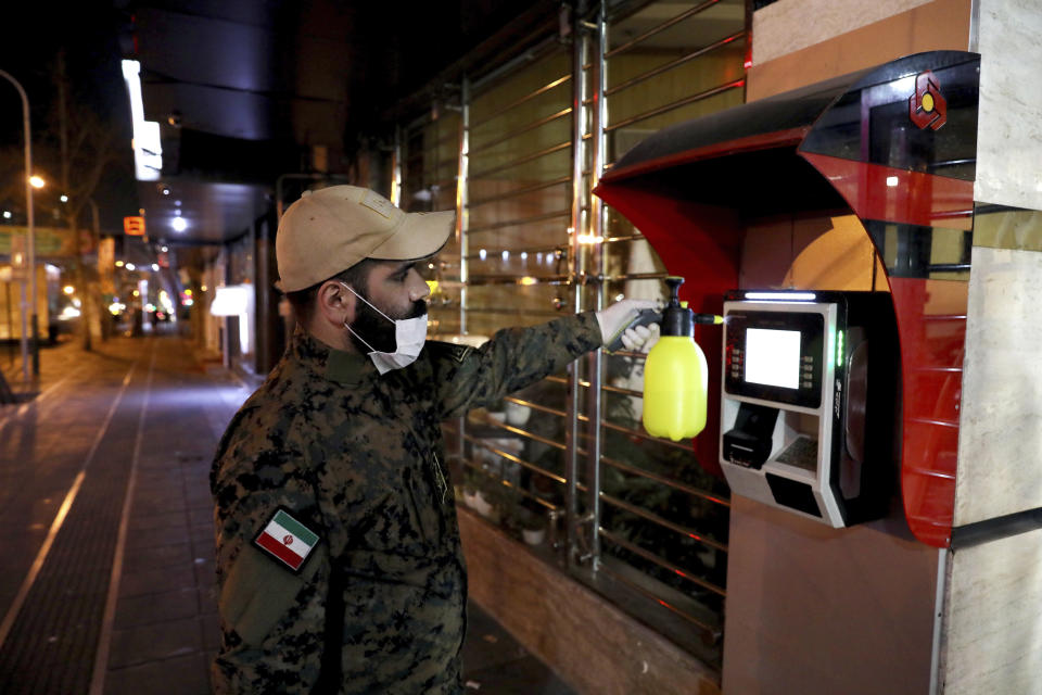 In this Wednesday, March. 4, 2020 photo, a Revolutionary Guard member disinfects an ATM machine to help prevent the spread of the new coronavirus in Tehran, Iran. Wearing gas masks and waterproof fatigues, members of Iran's Revolutionary Guard now spray down streets and hospitals with disinfectants as the Islamic Republic faces one of the world's worst outbreaks of the new coronavirus. (AP Photo/Ebrahim Noroozi)