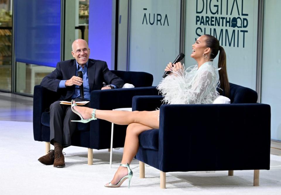 Chrissy Teigen in a chair onstage, kicking one leg out in front of her