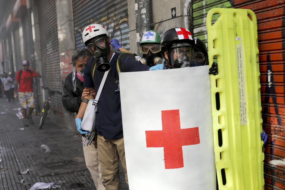 Paramedics, holding a shield imprinted with a red cross seal and a basket stretcher, standby as they watch clashes between anti-government demonstrators and police, in Santiago, Chile, Tuesday, Oct. 29, 2019. Chileans gathered Tuesday for a 12th day of demonstrations that began with youth protests over a subway fare hike and have become a national movement demanding greater socio-economic equality and better public services in a country long seen as an economic success story. (AP Photo/Rodrigo Abd)