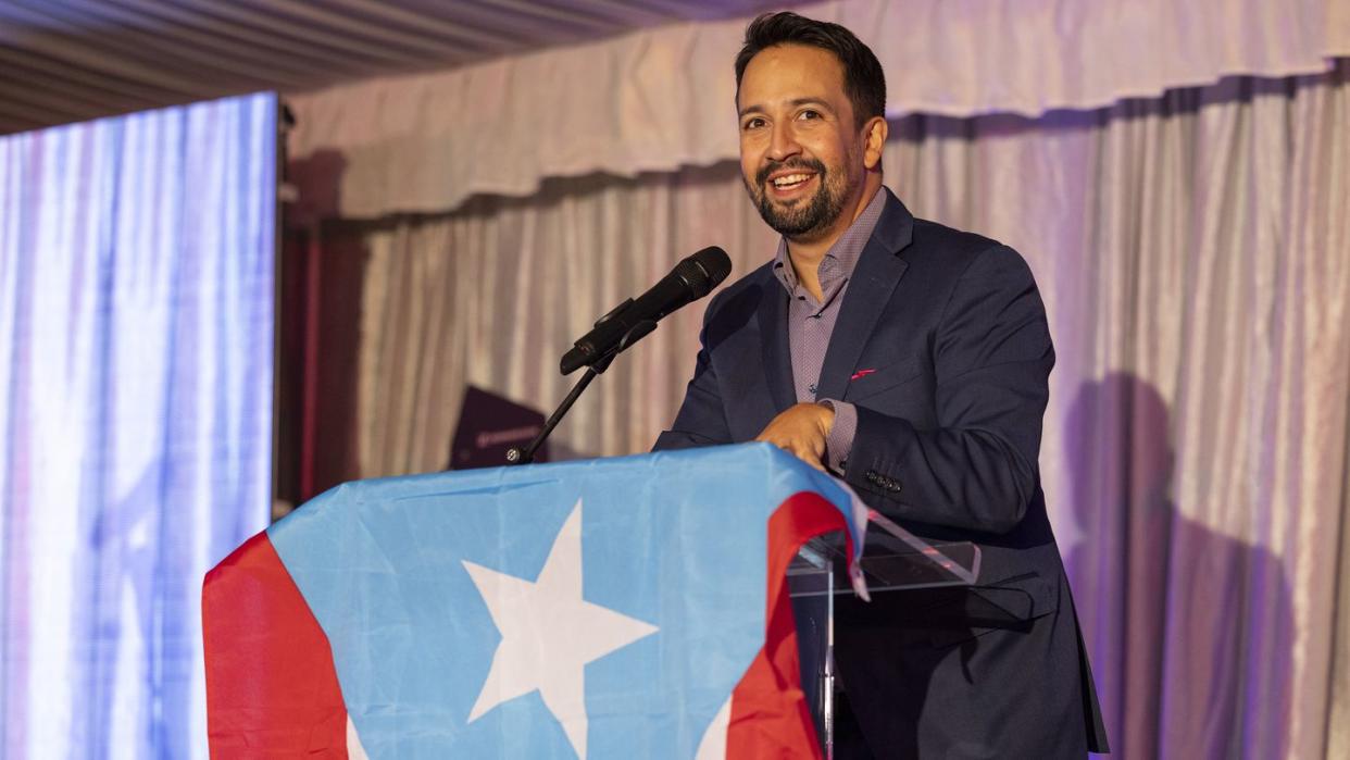 lin manuel miranda smiles and stands behind a podium draped with a puerto rican flag, he wears a dark suit with a purple unbuttoned collared shirt