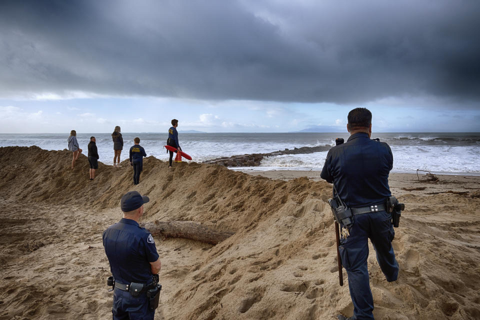 Police, life guards and spectators stand atop a sand berm and watch as turbulent surf hits the California coast on Saturday, Dec. 30, 2023 in Ventura, Calif. Bulldozers built giant sand berms to protect beachfront homes in Ventura, one of California's coastal cities hit hard this week by extraordinary waves generated by powerful swells from Pacific storms. (AP Photo/Richard Vogel)