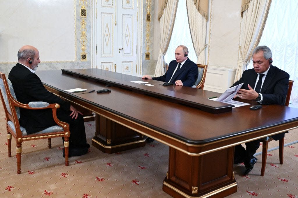 Putin at a long table talking to Iran's leader while Shoigu looks in a folder