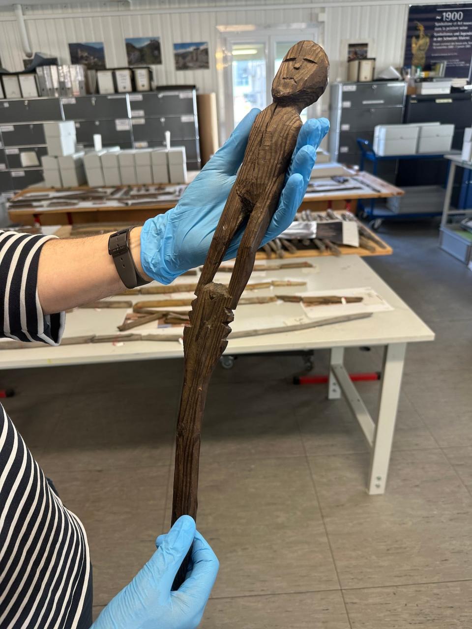 Blaue behandschuhte Hände halten eine kleine Holzskulptur eines Menschen mit flacher Miene und ohne Arme auf einem langen Holzpfahl