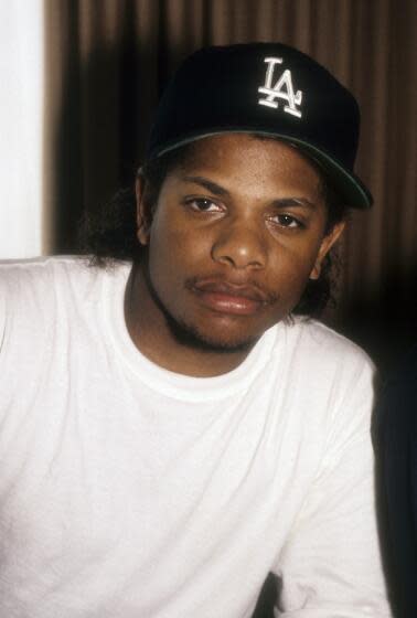 Eazy-E poses in a white t-shirt and a blue Dodgers "LA" baseball cap with a serious face