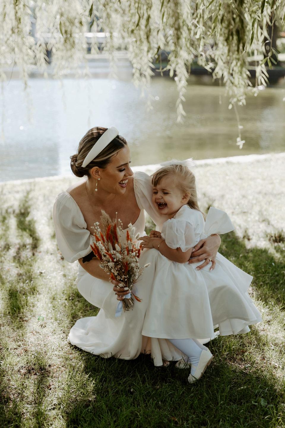 A bride kneels down to hug her flower girl in front of a lake.