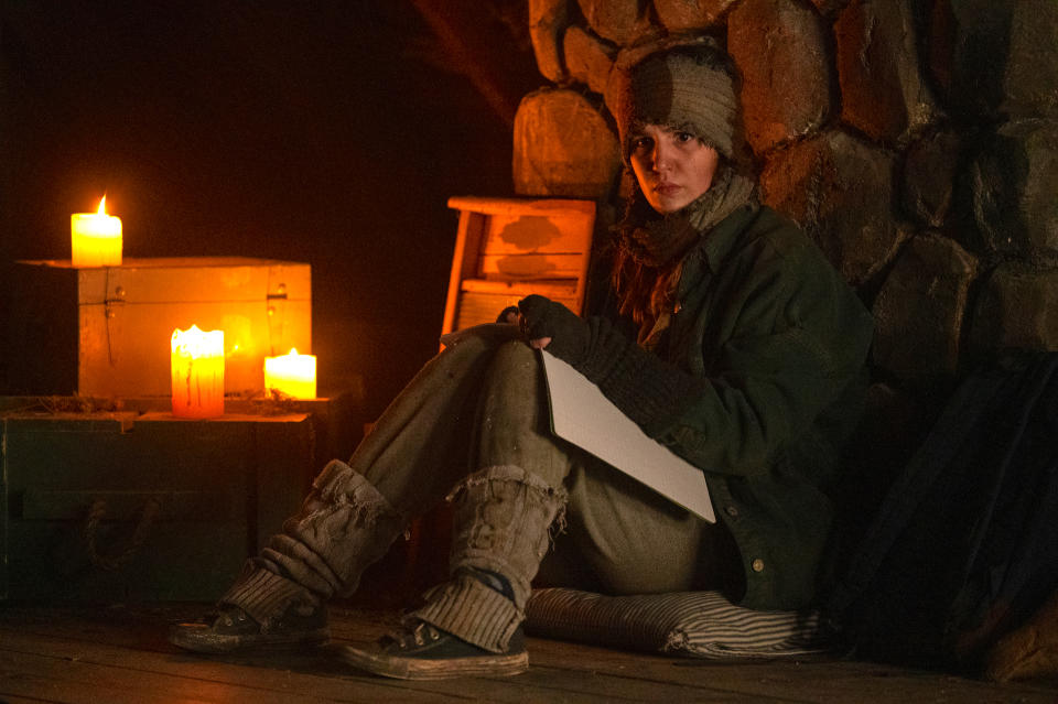 A teen girl bundled up in layered clothing, writing in a journal by candlelight; still from "Yellowjackets"