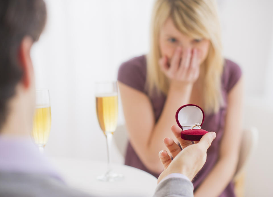 Man proposing to a woman with a ring, both seated with champagne glasses on table. Woman is emotionally covering her face