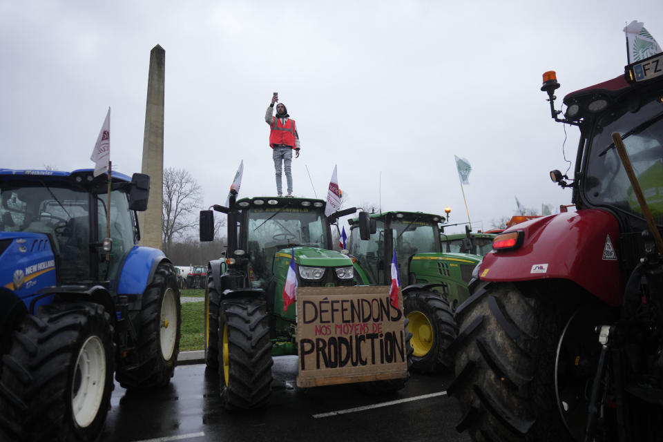 A farmer stands atop a tractor as farmers block a roundabout in Fontainebleau, south of Paris, Friday, Jan. 26, 2024 in Paris. Snowballing protests by French farmers crept closer to Paris with tractors driving in convoys and blocking roads in many regions of the country to ratchet up pressure for government measures to protect the influential agricultural sector from foreign competition, red tape, rising costs and poverty-levels of pay for the worst-off producers. Poster reads: Defend our production. (AP Photo/Thibault Camus)