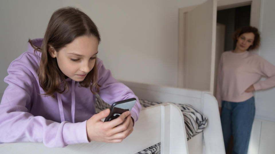 A young girl in a hoodie looks at her phone while leaning on a bunk bed; a woman stands in the doorway watching her