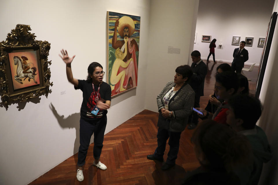 A guide explains to visitors about a painting showing 1910-17 Mexican revolutionary hero Emiliano Zapata nude, wearing high heels and a pink, broad-brimmed hat, straddling a horse, in Mexico City, Tuesday, Dec. 10, 2019. The work by Fabian Chairez is part of an exhibit about Zapata in one of Mexico City's premiere arts venues, the Fine Arts Palace. (AP Photo/Eduardo Verdugo)