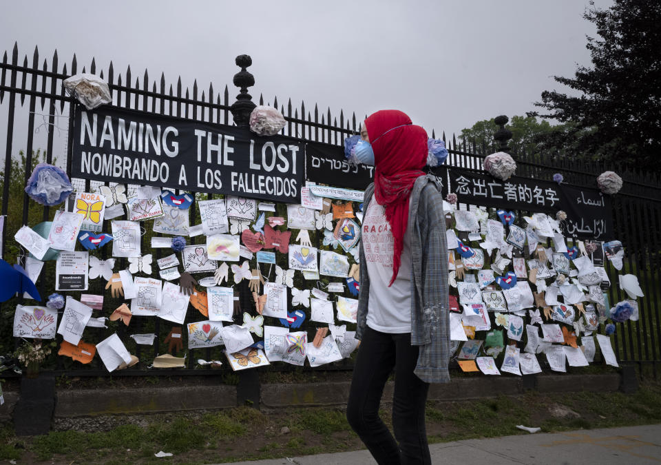 A woman passes a fence outside Brooklyn's Green-Wood Cemetery adorned with tributes to victims of COVID-19, Thursday, May 28, 2020, in New York. The memorial is part of the Naming the Lost project which attempts to humanize the victims who are often just listed as statistics. The wall features banners that say "Naming the Lost" in six languages: English, Spanish, Mandarin, Arabic, Yiddish and Bengali. (AP Photo/Mark Lennihan)