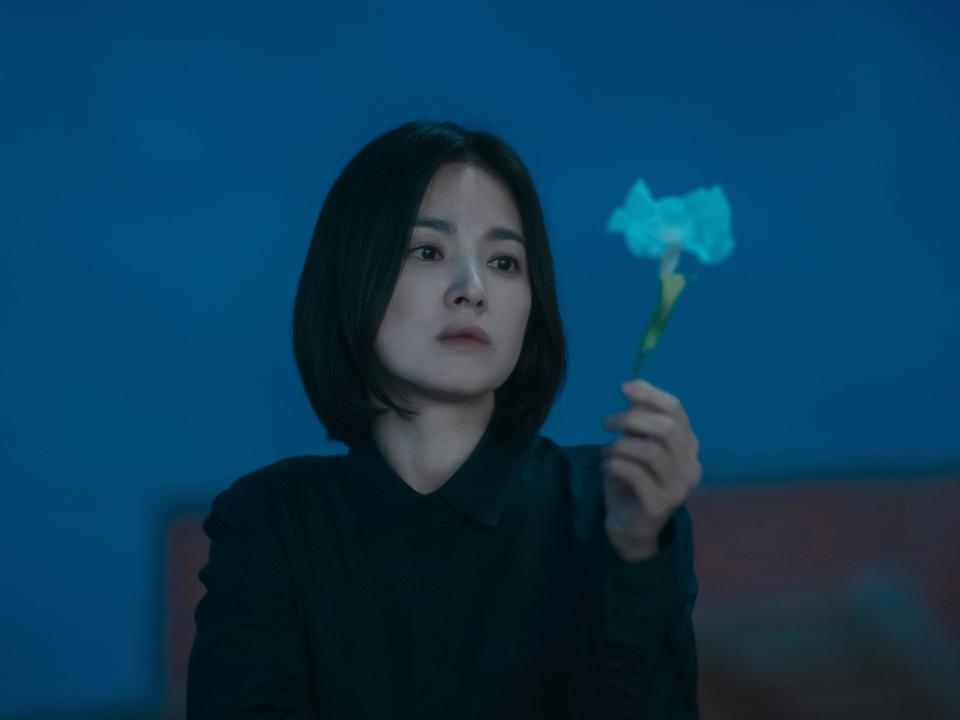 song hye-kyo as moon dong-eun in the glory, a woman in her 30s with a severely cut bob, standing on a rooftop at disk and holding a white flower out in front of her