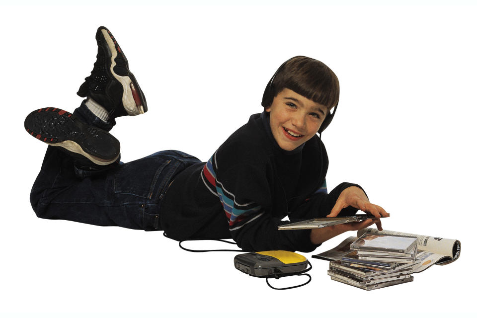 A '90s kids lying on the floor listening to a CD from his portable CD player while he has a CD case opened in his hand and has a stack of CDs next to him