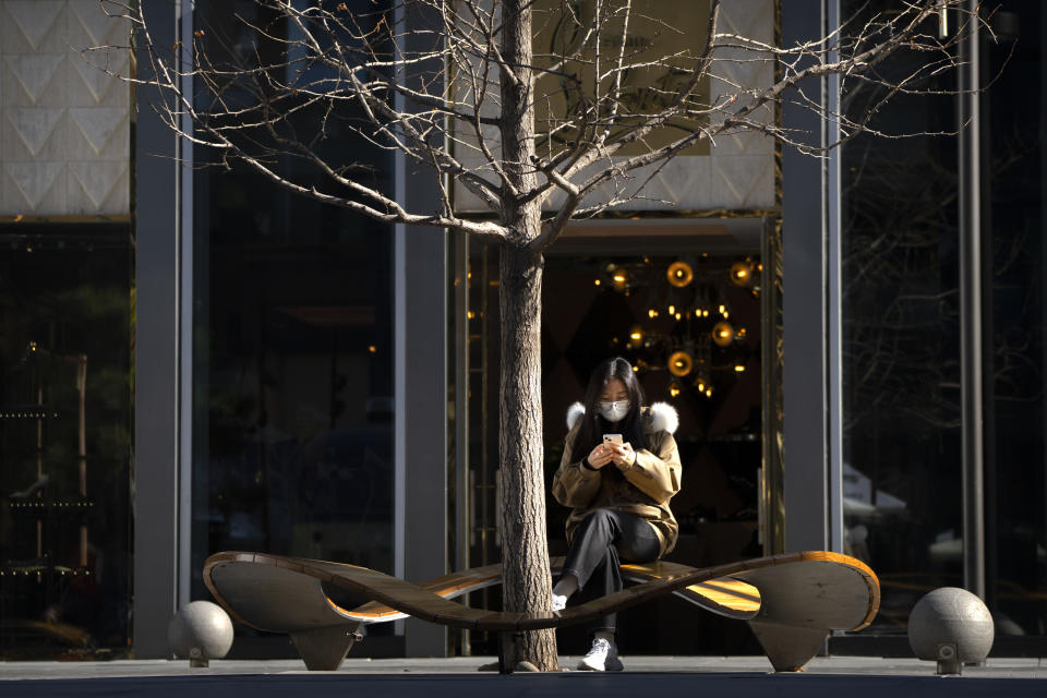 A woman wearing a face mask sits on a bench at an outdoor shopping complex in Beijing, Saturday, Nov. 5, 2022. (AP Photo/Mark Schiefelbein)