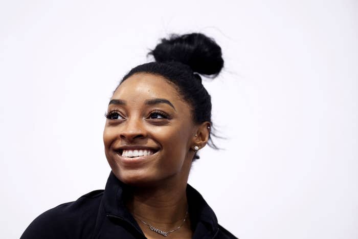 Simone Biles smiling, wearing a jacket and a necklace, with her hair styled in a bun