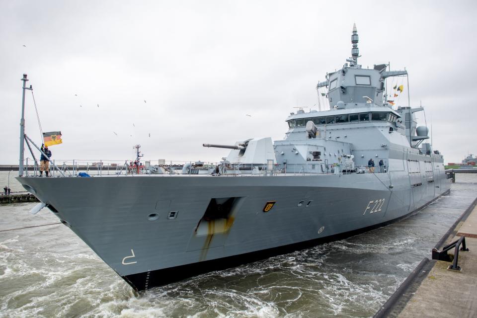 The front-left of the German Navy frigate Baden-Württemberg at port, with churning waters below and a gray sky above.