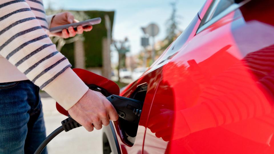 Horizontal view of cropped male using phone while charging red electric car battery at residential street.