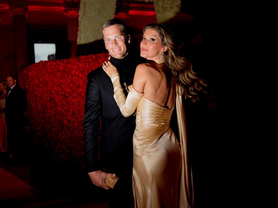 Tom Brady in black tux and and Gisele Bündchen in gold dress in front of red decor