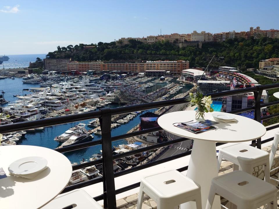 Ein Balkon mit schwarzem Geländer und weißen Stühlen und Tischen mit kleinen Blumensträußen darauf. Die französische Riviera und mehrere Hotels, Bäume und andere Gebäude sind im Hintergrund zu sehen.