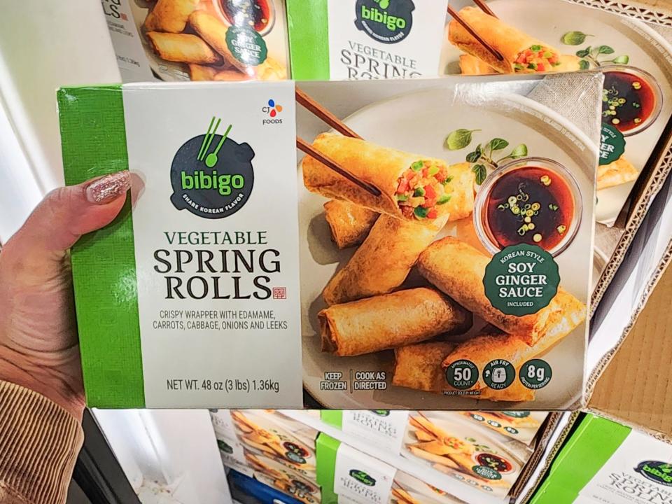 A hand holds a box of Bibigo vegetable spring rolls with images of spring rolls and soy sauce on the box