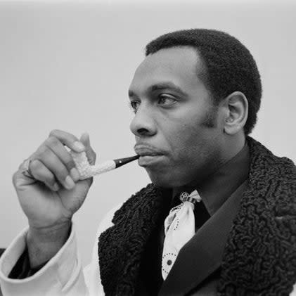 American guitarist and singer Freddie Roulette, circa 1970, wearing a white and black textured jacket, holding a pipe near his mouth, looking contemplative to the side