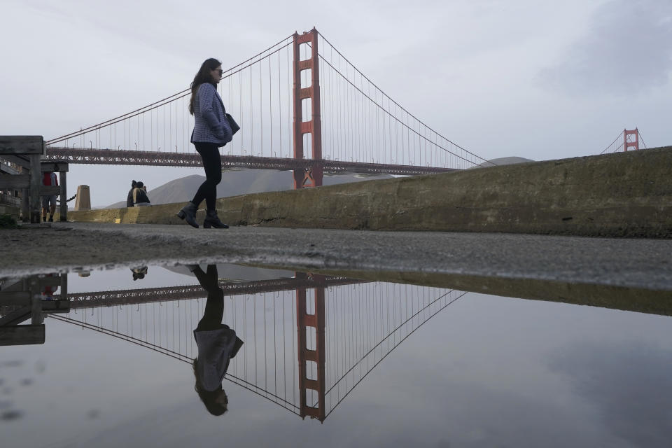 A pedestrian and the Golden Gate Bridge are reflected in a puddle on a path near Crissy Field in San Francisco, Friday, Jan. 6, 2023. California weather calmed Friday but the lull was expected to be brief as more Pacific storms lined up to blast into the state, where successive powerful weather systems have knocked out power to thousands, battered the coastline, flooded streets, toppled trees and caused at least six deaths. (AP Photo/Jeff Chiu)