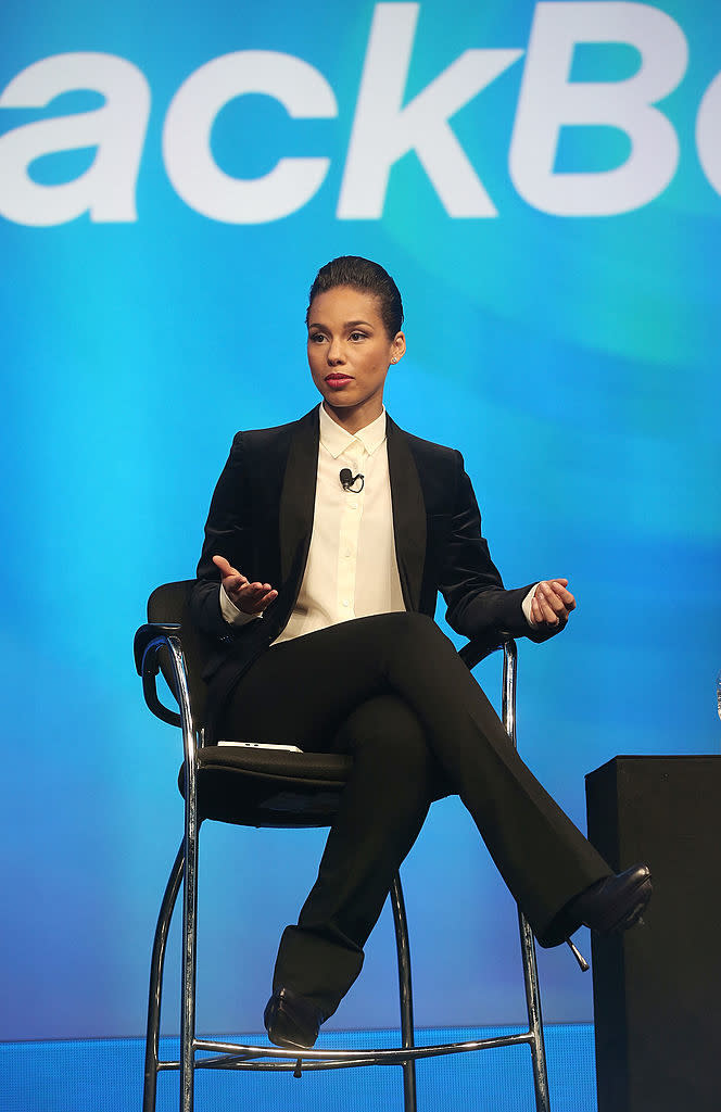 Alicia Keys sits on stage, dressed in a sleek black suit with a white shirt, during a speaking engagement