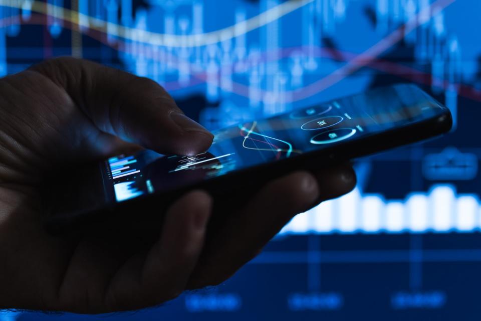 A hand holds a smartphone displaying stock charts.