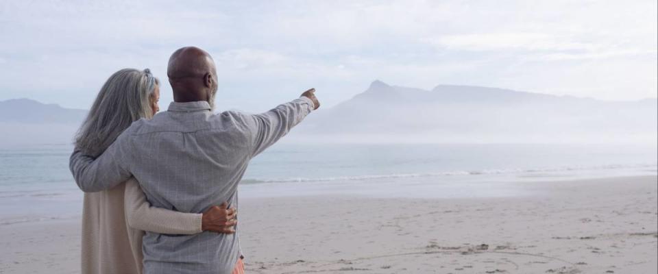 Rear view of happy senior African-American couple standing next to each other while looking at the sea and mountains on beach on beautiful cloudy day.