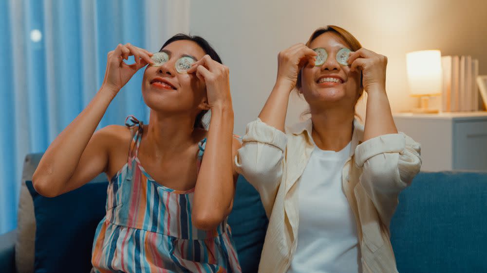 Young Asian female lesbian in living room. Happy girls having fun use daily cucumber eyes mask patch together sit on cozy sofa couch at night.