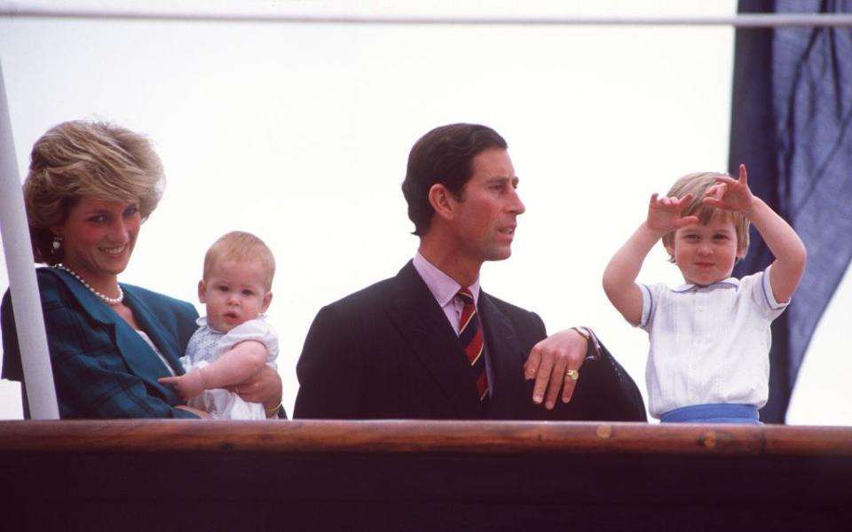 princess diana holds baby prince harry and stands next to then prince charles and prince william behind a wooden railing