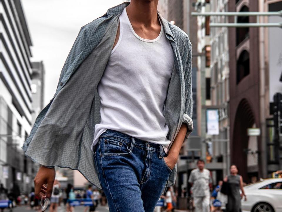 man walking down a street wearing a white tank top and a gray over shirt with a pair of jeans