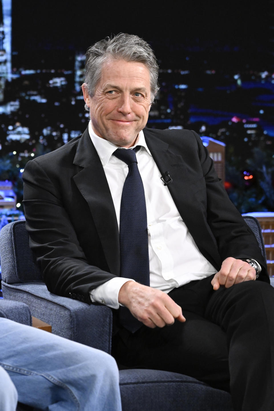 Hugh Grant in a suit and tie, smiling while seated during a talk show appearance