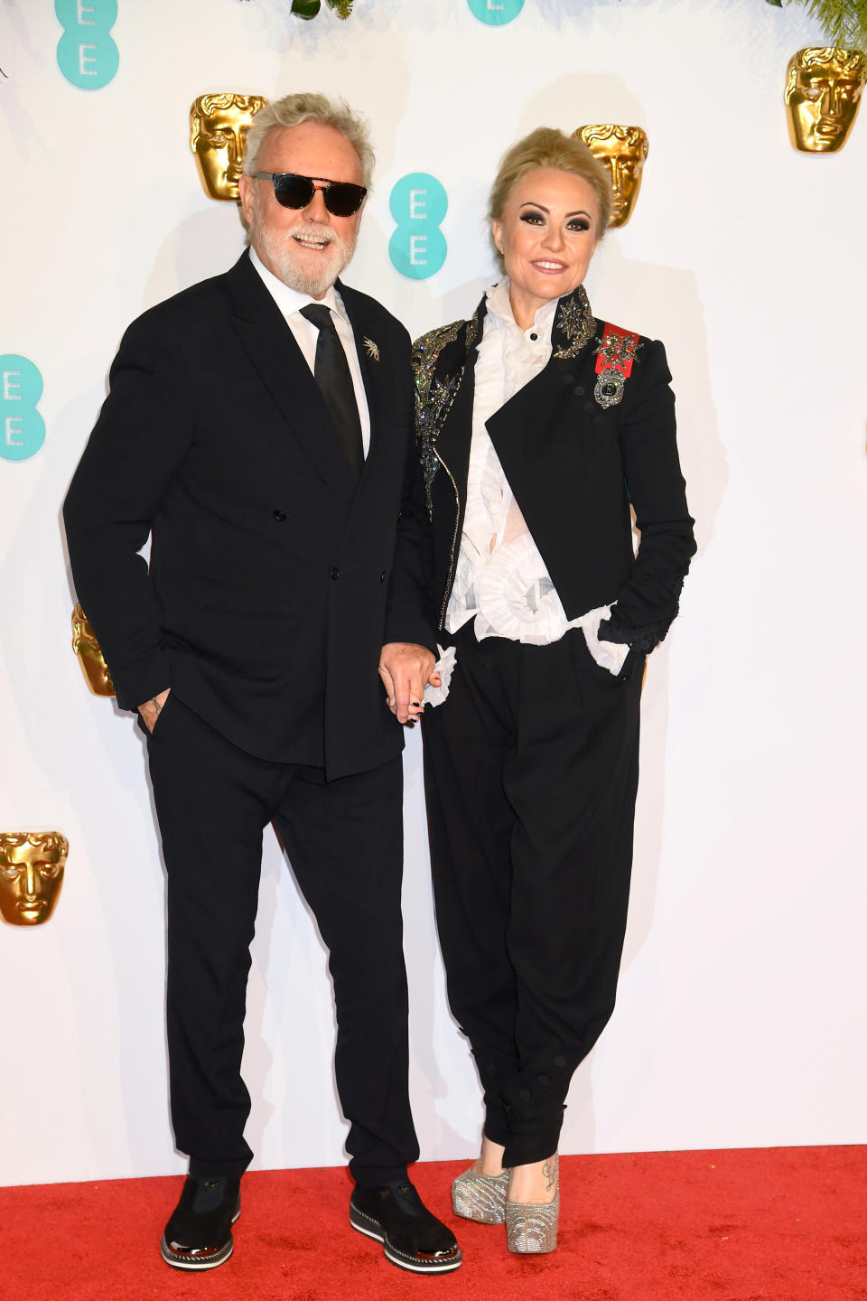 Roger Taylor and Sarina Potgieter attend the EE British Academy Film Awards. Sarina wears a frilly black blouse, a black blazer with military style badges in red and gold, baggy trousers and golden platform heels.