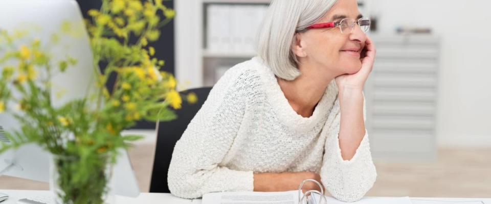 Happy Thoughtful Senior Businesswoman Sitting at her Table Inside the Office and Looking Into the Distance.