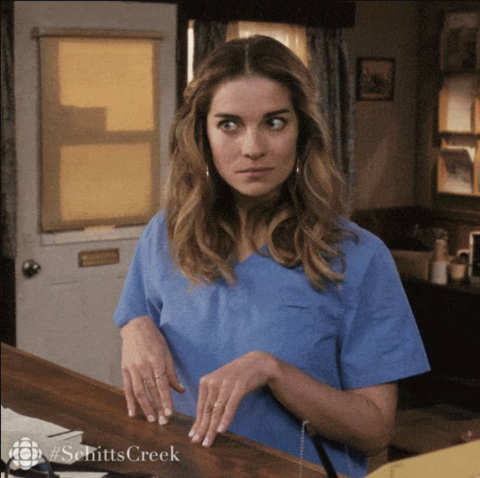 Annie Murphy as Alexis Rose from Schitt's Creek, wearing a blue shirt, makes an unsure facial expression with her hands poised in front of her. #SchittsCreek