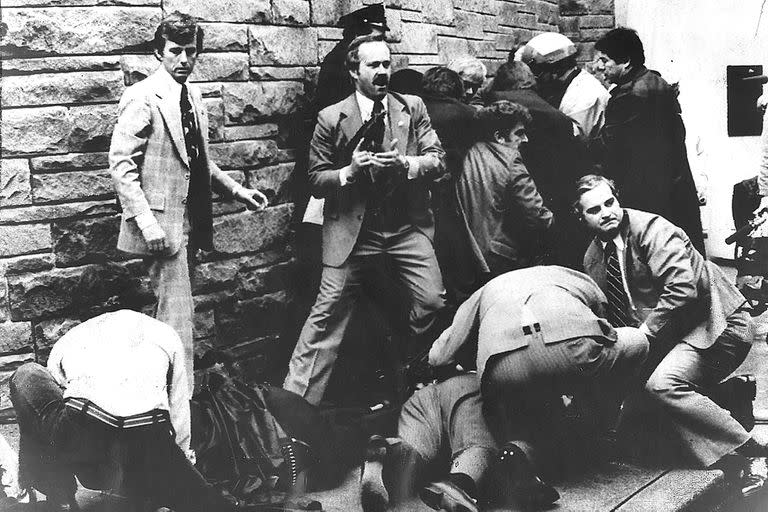 AGENTS TEND TO PRESIDENTIAL PRESS SECY JAMES BRADY ON THE GROUND AR RIGHT AND A POLICEMEN (LEFT) WHO WERE WOUNDED 3/30 IN AN ASSASINATION ATTEMPT ON PRESIDENT REAGAN. THE ASSAILANT IS BEING HELD BY POLICE AND AGENTS IN BACKGROUND (RIGHT). 3/30/81